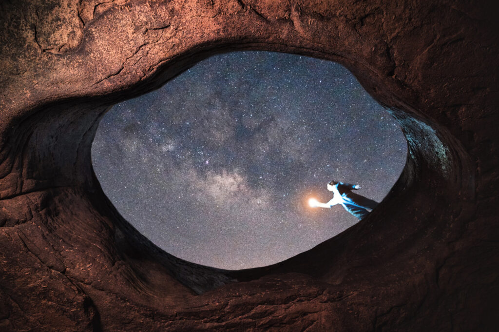 Pessoa com lanterna na entrada de uma grande caverna sob a Via Láctea, ilustrando a busca por vida alienígena no subsolo